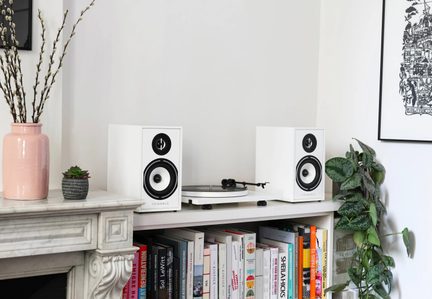 Par de caixas de som de estante brancas em material rígido, gabinetes retangulares vistos de frente com tweeter e woofer pretos com bordas, posicionadas de cada lado de um toca-discos em uma prateleira