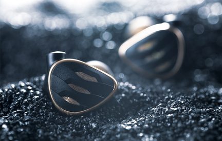 Dreiviertel-Nahaufnahme von zwei dreieckigen In-Ear-Kopfhörern mit mattschwarzem Gehäuse und goldmetallischer, durchbrochener Zierleiste, liegend auf einer dunklen, körnigen Oberfläche
