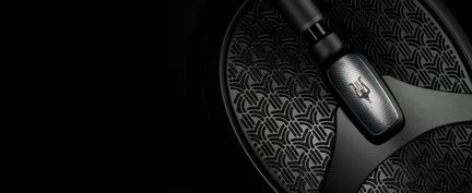 Tæt, skråt billede af en sort, tekstureret metalcirkel med et geometrisk vifteformet mønster, gennemskåret af en mat arm, der er udsmykket med en børstet metalplade med et tretakket symbol.