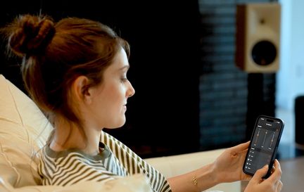 Smartphone mit Audio-Einstellungs-App in der Hand einer Frau auf dem Sofa, vernetzter Lautsprecher im Hintergrund