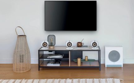 Sort vægmonteret fjernsyn, lavt møbel i træ og metal, 4 hvide højttalere, gitterlampe, brun vase, hvid kasse, flettet kurv