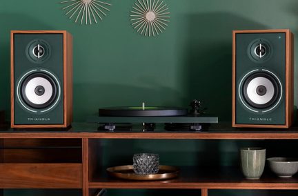 Două boxe Triangle pe un mobilier de lemn, un pick-up negru între boxe, obiecte decorative pe perete