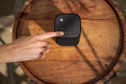 Mano che tiene un altoparlante intelligente nero rotondo visto frontalmente su un vassoio di legno, primo piano