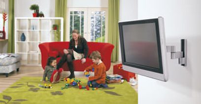 Billedet viser en stue med en mand, der sidder på en rød sofa og leger med to små børn på et grønt tæppe med legetøj spredt ud, foran et stort fladskærms-tv, der er monteret på væggen