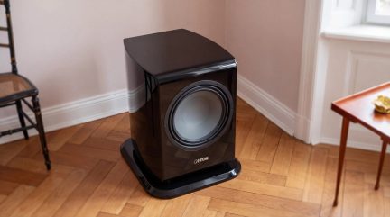 Canton subwoofer i blank svart med grå frontmontert høyttaler, plassert på gulvet i et hjørne av rommet