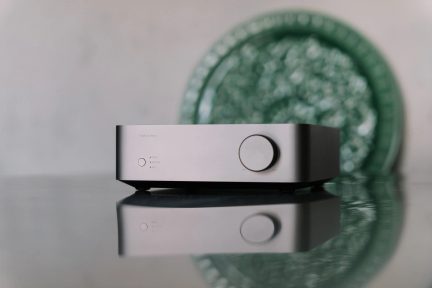 Amplificador de áudio compacto cinza com grande botão de volume em vista frontal sobre superfície refletora
