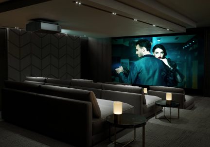 Sala de cine privada con sofás grises, mesas redondas negras, lámparas cilíndricas, pantalla gigante, proyector en el techo