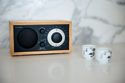 Radio Bluetooth Tivoli Audio Model One BT iz svetlega lesa, s črno sprednjo ploščo in velikim okroglim gumbom, postavljen na beli mizi