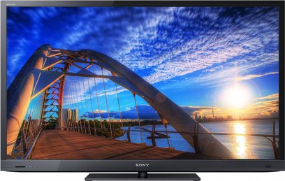 Sony KDL-46EX720 tv set forfra, tændt skærm der viser en buet metalbro over vandet ved solnedgang, blå himmel med skyer