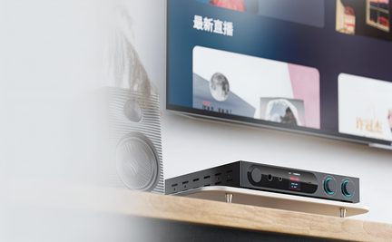 Amplificatore audio rettangolare nero in metallo, visto da tre quarti davanti, pannello frontale con due grandi manopole retroilluminate blu, piccolo schermo centrale a colori, prese circolari e fessure di ventilazione laterali