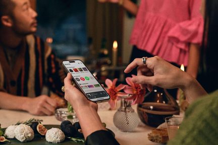 Hånd med ringer, sølvfarget smarttelefon i metall, skjerm med musikkapp, sett ovenfra et bord med desserter og blomster