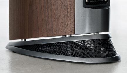 Nærbillede i frøperspektiv af den elliptiske, perforerede sokkel på en gulvhøjttaler, der kombinerer åretegnet valnødfinér og satin-gråt metal