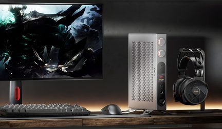 Postazione informatica scura con ampio schermo che mostra una scena di gioco, piccolo case verticale argentato in metallo traforato con due manopole luminose e frontale nero, tastiera meccanica nera, mouse nero e cuffie nere su supporto, vista frontale