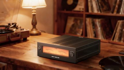 FiiO R2R Tube DAC sort audiokonverter, set fra trekvartsvinkel med baggrundsbelyst VU-meter på bord nær en vinylpladespiller