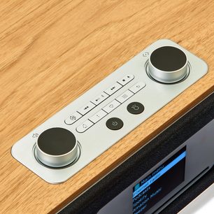Ovenfra set i let perspektiv, Roberts Stream 67 bordradio i lys træfinér med kontrolpanel i børstet aluminium, to store sølvfarvede drejeknapper med sorte toppe, der flankerer en central række rektangulære knapper og to runde sorte knapper.