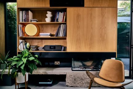 Diffusore audio nero con schermo centrale su una mensola, in un soggiorno con camino e poltrona color cammello