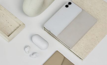 Bovenaanzicht van een witte smartphone met een matte, gladde achterkant en een dubbele zwarte cameramodule, een wit draadloos oplaadetui en twee bijpassende oordopjes, geplaatst op notitieboekjes en rechthoekige dienbladen van beige stof en licht steen.