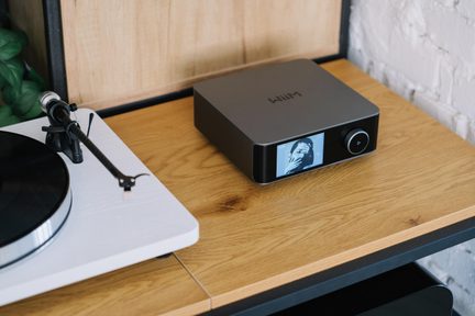 Hi-fi veriga, sestavljena iz antracitno sive kovinske avdio enote, prikazane v rahlo poševnem pogledu od spredaj, s sijočo črno sprednjo ploščo, v katero sta vgrajena barvni zaslon in okrogla vrtljiva tipka, postavljena desno od bele gramofonske plošče s črnim krožnikom na leseni polici.