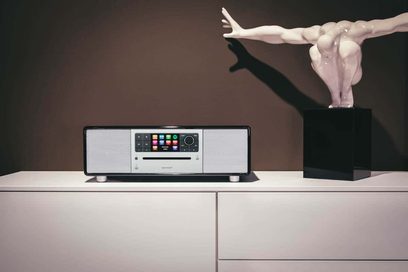 Sonoro Prestige sort radioboks, blank sort og sølv, set forfra, stående på hvidt møbel. Hvid skulpturel arm oven over