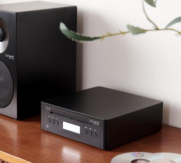Zwarte cd-speler van mat metaal met sleuf, lcd-scherm en ronde knoppen, driekwart vooraanzicht op een houten meubel