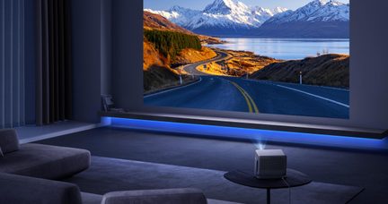 Dunkles Wohnzimmer mit weißem Kunststoffprojektor auf rundem, schwarzem Couchtisch, Leinwand projiziert Straßenlandschaft und Berge