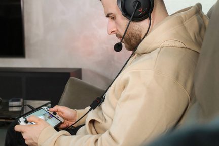 Person in profile, black plastic over-ear headset with boom microphone, beige sweatshirt, white plastic handheld console