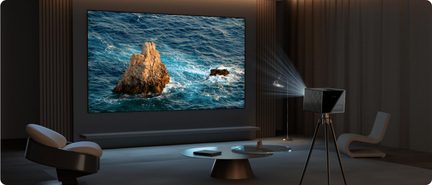 Ribbed black metal video projector mounted on a metal tripod, three-quarter view, light beam directed toward a large screen