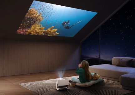 Barn sidder på pude med bamse, kigger på undervandsprojektion i loftet, projektor på tæppe, sofa til højre