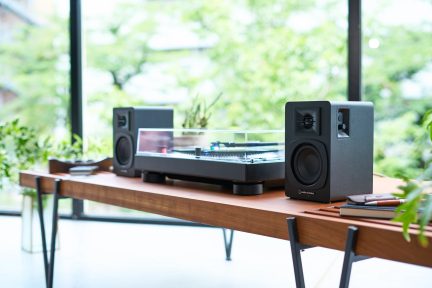 Dřevěný stůl s černou vinylovou deskou pod průhledným krytem, dvě černé reproduktory, boční pohled, rozmazané pozadí