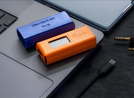 Due piccoli dispositivi rettangolari in plastica blu e arancione con superficie testurizzata, ciascuno dotato di una finestra per lo schermo e di fori laterali, visti in primo piano con una leggera inquadratura dall’alto, posizionati sull’angolo di un computer portatile grigio