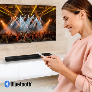 Smilende kvinde, der holder en smartphone og kigger på et TV-skærm, der viser en menneskemængde til en koncert, Bluetooth-teknologi