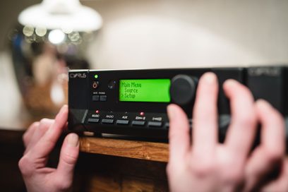 Amplificador hi-fi estéreo Cyrus 6 DAC QXR, vista aproximada da frente com visor verde "Menu Principal Configuração de Fonte", mãos nos botões
