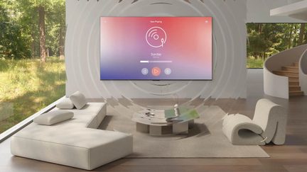 Pohled zepředu na minimalistický obývací pokoj v světlých tónech s nízkou modulární pohovkou v béžové látce, zakřiveným šedým látkovým křeslem, kulatým kovovým stolkem s leštěným povrchem a černou plochou televizí na stěně zobrazující růžovo-fialový přechod, zadní stěna s reliéfními koncentrickými vzory a prosklená stěna s výhledem do lesa.