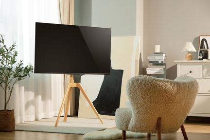 Sort glasfladskærms-tv monteret på et lyst træstativ i staffeli-stil, set forfra i en lys stue