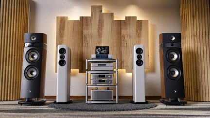 Ezüst színű fém polcokból álló hi-fi rendszer frontális nézetben, amelyen lemezjátszó és erősítők helyezkednek el, tetején egy táblával. Két oldalán két-két fehér, karcsú, kör alapú oszlophangszóró és két fekete, fa oldallapokkal rendelkező, szélesebb oszlophangszóró áll. Az egész szőnyegen helyezkedik el egy fa lamellás akusztikai panel előtt.