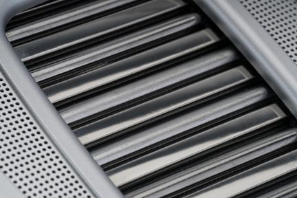 Nært billede i skrå vinkel af en grå metalventilationsrist med blanke lameller og perforerede paneler