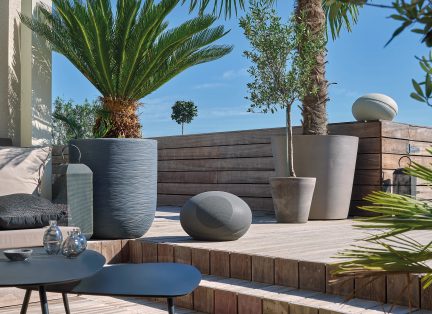 Grijzen keramische potten en vazen in verschillende vormen op een houten terras met palmbomen en een blauwe lucht op de achtergrond