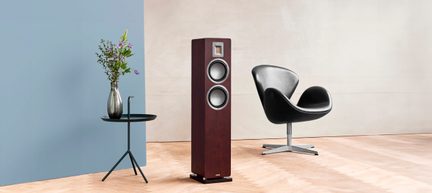 Interno minimalista, diffusore a colonna in legno marrone scuro con due altoparlanti circolari argentati e tweeter rettangolare sul frontale, vista frontale, posizionato su parquet chiaro, accanto a un tavolo rotondo nero con un vaso di fiori e a una poltrona girevole nera