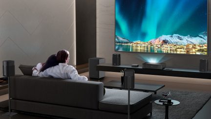 Bagudsigt af en moderne stue med grå stofsofa, stor vægmonteret skærm, der viser nordlys, sorte surroundhøjttalere i metal og plast, kompakt projektor og subwoofer placeret på sort hylde