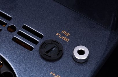 Detailní šikmý záběr na horní černý kovový texturovaný panel trubkového zesilovače Cayin CS-805A, který ukazuje ventilační štěrbiny, oranžový nápis „A2 FUSE“, černou kruhovou pojistkovou zátku a stříbrný závitový vložku.