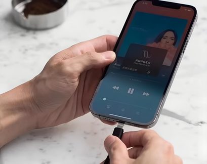 Sort opladerkabel tilsluttet en iPhone, der afspiller musik, close-up af hænder på marmoreret bordbaggrund