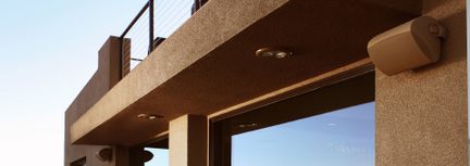 Beżowa fasada kątowa z metalową balustradą, głośnikami ściennymi, wbudowanymi reflektorami, dużymi przeszklonymi oknami odbijającymi światło