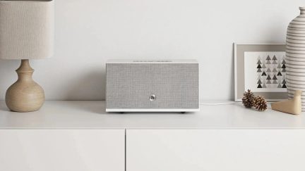 Coluna sem fios retangular cinzenta com frente texturizada e controlos na parte superior, colocada sobre um móvel branco