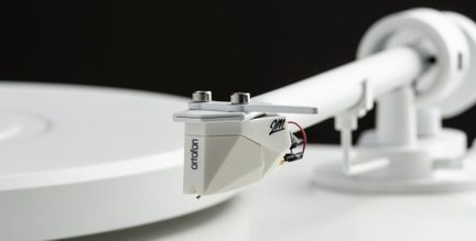 Nærbillede af den matte hvide plastik-pickup med sort logo, fastgjort med to sølvfarvede skruer på den hvide, rørformede metal-tonearm, set fra trekvart forfra