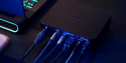 Mat sort aluminiums kabinet, set bagfra i trekvart vinkel, række af mærkede stik: coaxial, XLR, USB-B og strømforsyning med tilsluttede kabler, diskret logo centreret på toppen under blåt lys