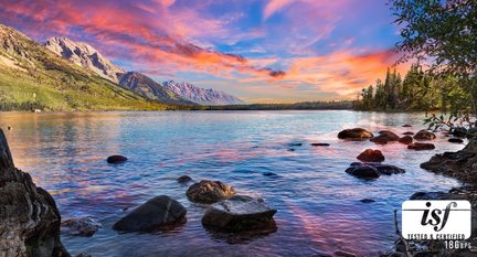 Lac albastru cu reflexii roz-portocalii, mărginit de stânci întunecate și copaci verzi, dominat în stânga de un lanț muntos verzui și gri sub un cer crepuscular multicolor, vedere frontală de pe mal