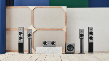 Sæt med seks hi-fi-højttalere i grå metal og sort træ, fronter i børstet aluminium med synlige runde højttalerenheder, bestående af to gulvhøjttalere, to reolhøjttalere på standere, en subwoofer og en centerhøjttaler, set forfra
