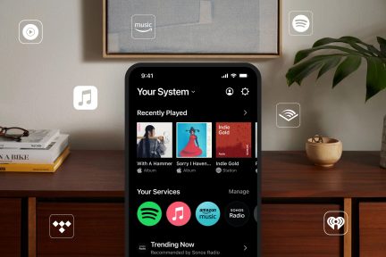 Smartphone con schermo nero che mostra applicazioni musicali, posato su un mobile in legno, occhiali su libri, pianta verde, ciotola beige