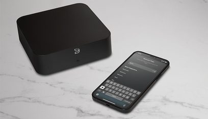 Cassa nera opaca quadrata e smartphone con app di controllo Bluesound, vista dall'alto su superficie marmorea grigia