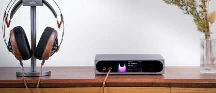 Taisnstūrveida pelēks alumīnija audio pastiprinātājs‑atskaņotājs, skats no priekšpuses, spīdīga melna priekšpuse ar krāsu ekrānu labajā pusē, blakus apļveida skārienpaliktnis un pa kreisi 6,35 mm austiņu ligzda, kas savienota ar vara pīto kabeli